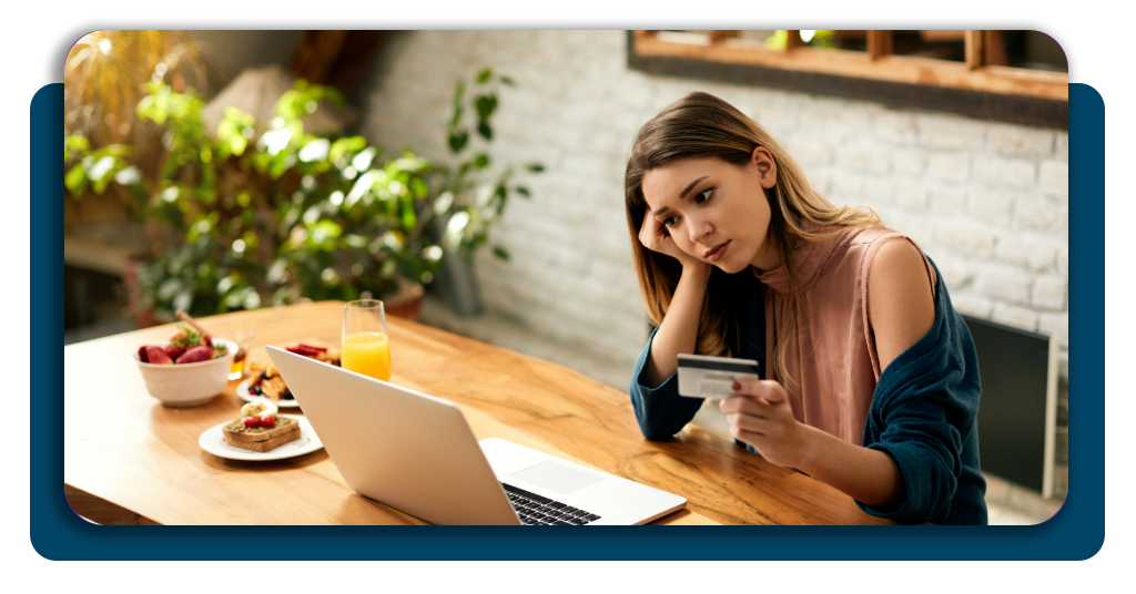 A young woman reviewing her credit card statements while sitting in front of a laptop at the kitchen table with a focused and worried expression.